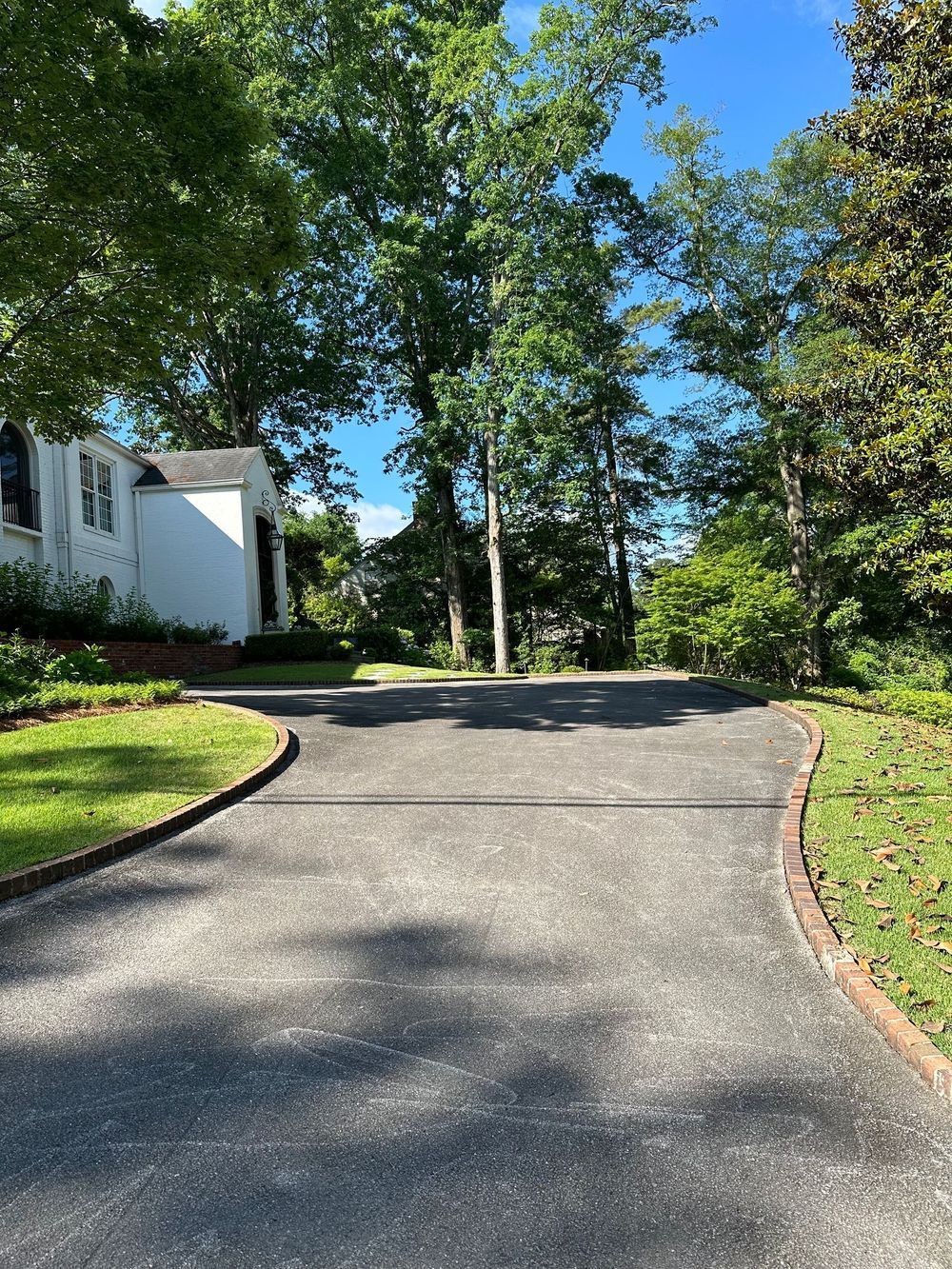 Driveway leading to a white house surrounded by trees. Green grass borders the asphalt.