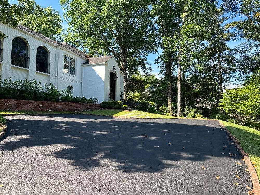 A paved driveway leads to a white building with arched windows. Lush greenery surrounds the scene on a sunny day.