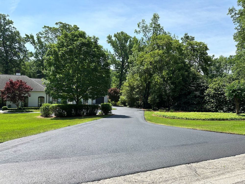 Paved driveway leading to a white house with green lawn and trees under a bright blue sky.