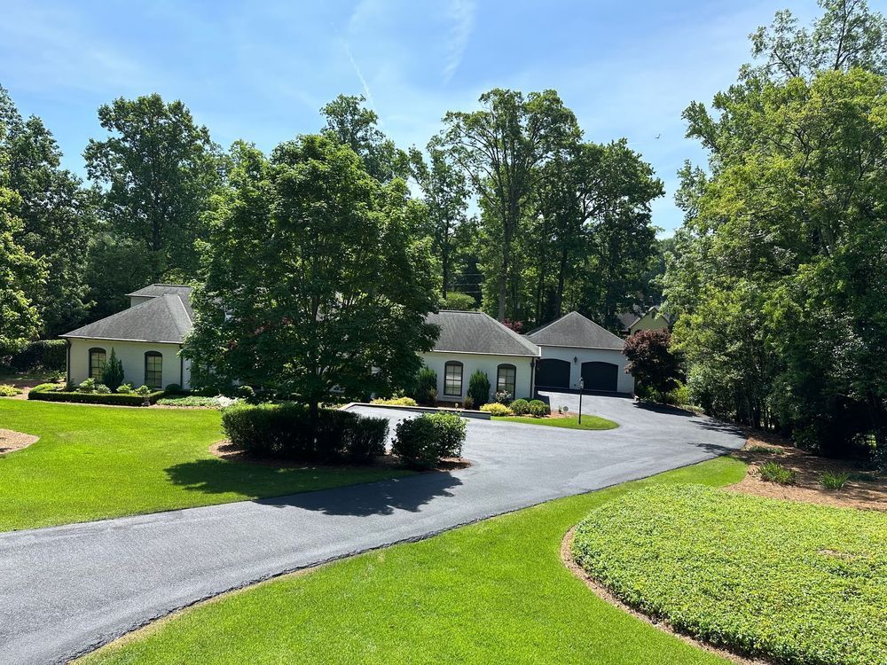 A large, light-colored house with a curved driveway and attached garage, surrounded by green grass and trees on a sunny day.