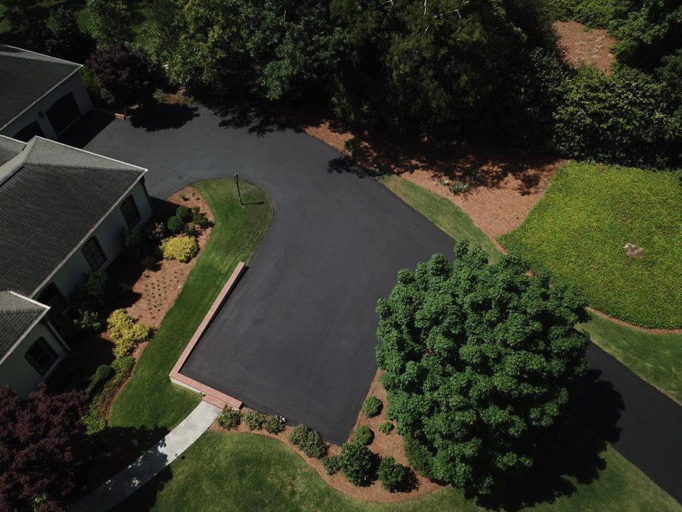 Aerial view of a newly paved black asphalt driveway winding past a brick wall, houses, and lush green trees.