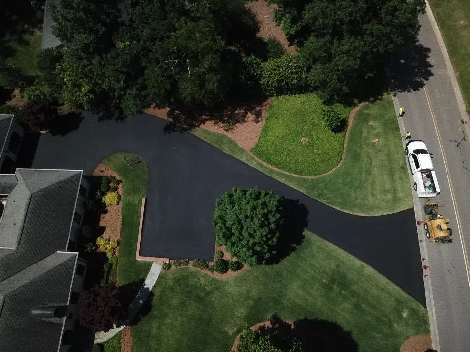 Overhead view of newly paved asphalt driveway winding past lawns and trees, next to a road with a white truck parked.