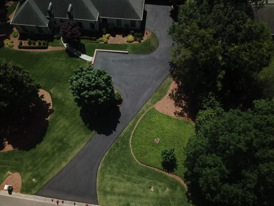 Aerial view of a house with a black asphalt driveway winding through green lawns and trees.