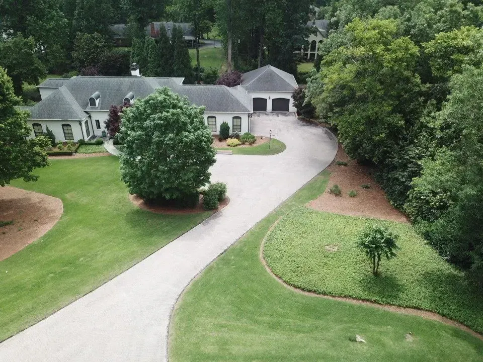 Aerial view of a house with a long driveway surrounded by green grass and trees. A two-car garage is attached.