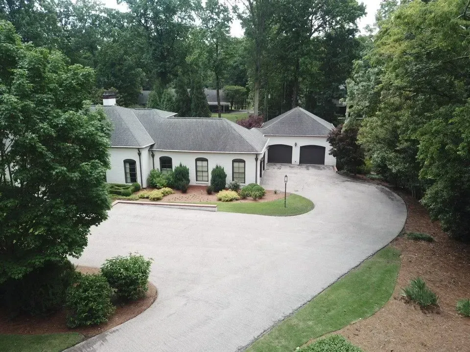 White house with black trim and a two-car garage, set amongst lush green trees with a long paved driveway.