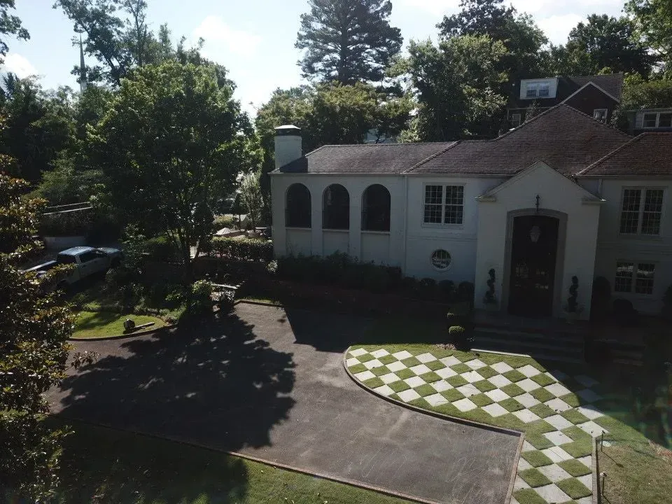 A white house with a checkered-patterned walkway and a dark asphalt driveway, surrounded by green trees.