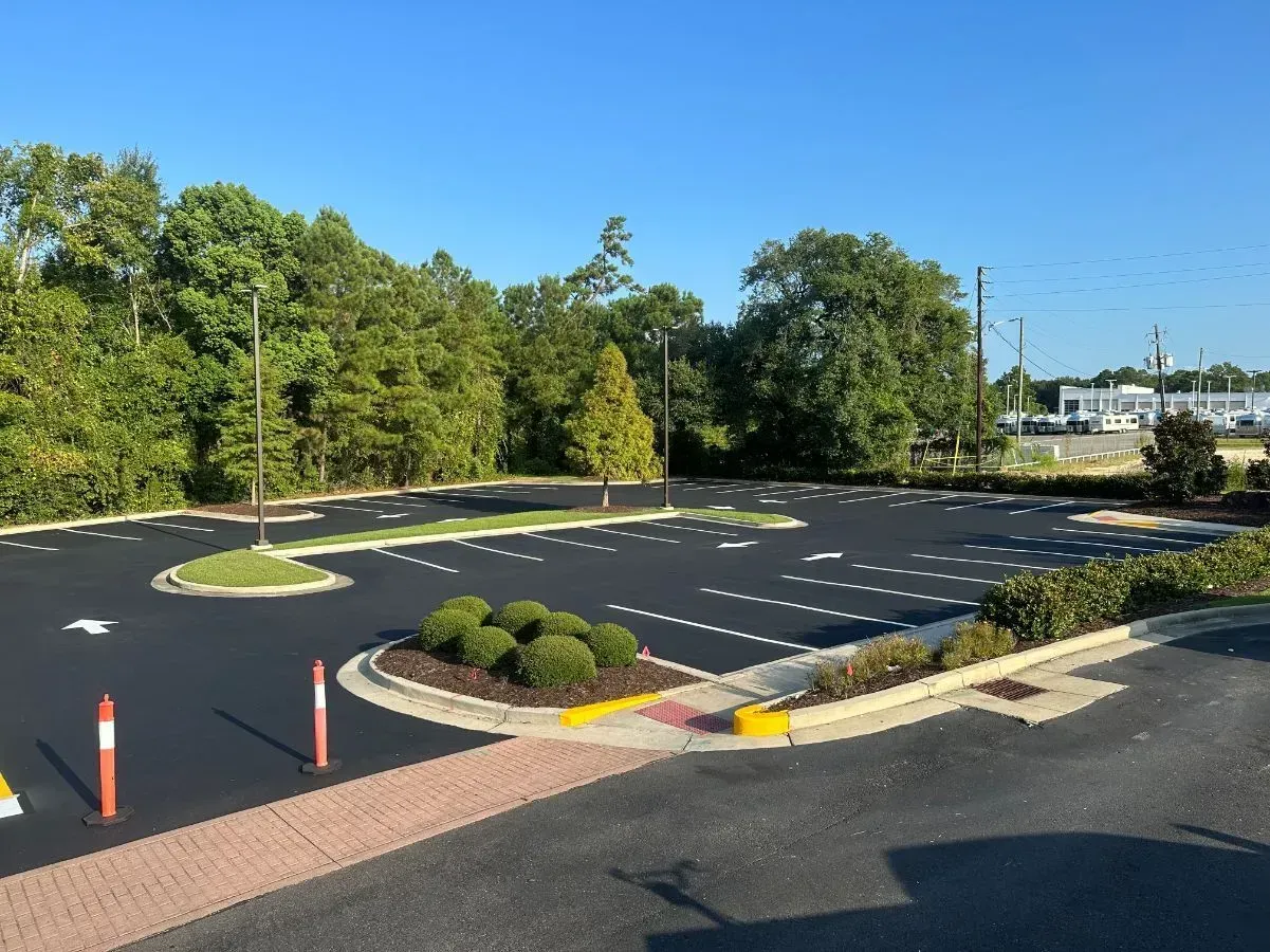 Empty asphalt parking lot bordered by landscaping and a brick-lined sidewalk, trees in the background, and blue sky.