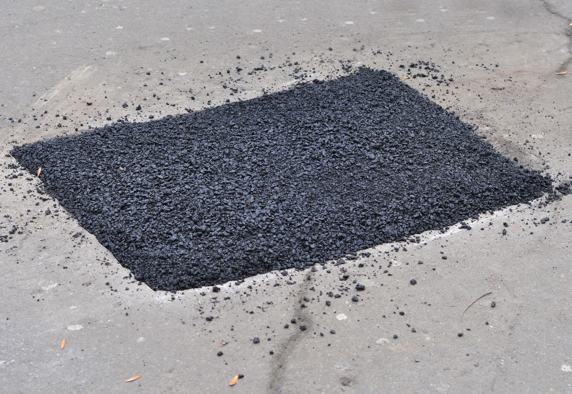 Square asphalt patch on a gray road surface, filled with dark, loose gravel.