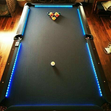A pool table in a room with hardwood floors, featuring a racked set of balls, a white cue ball, and glowing blue LED edges.