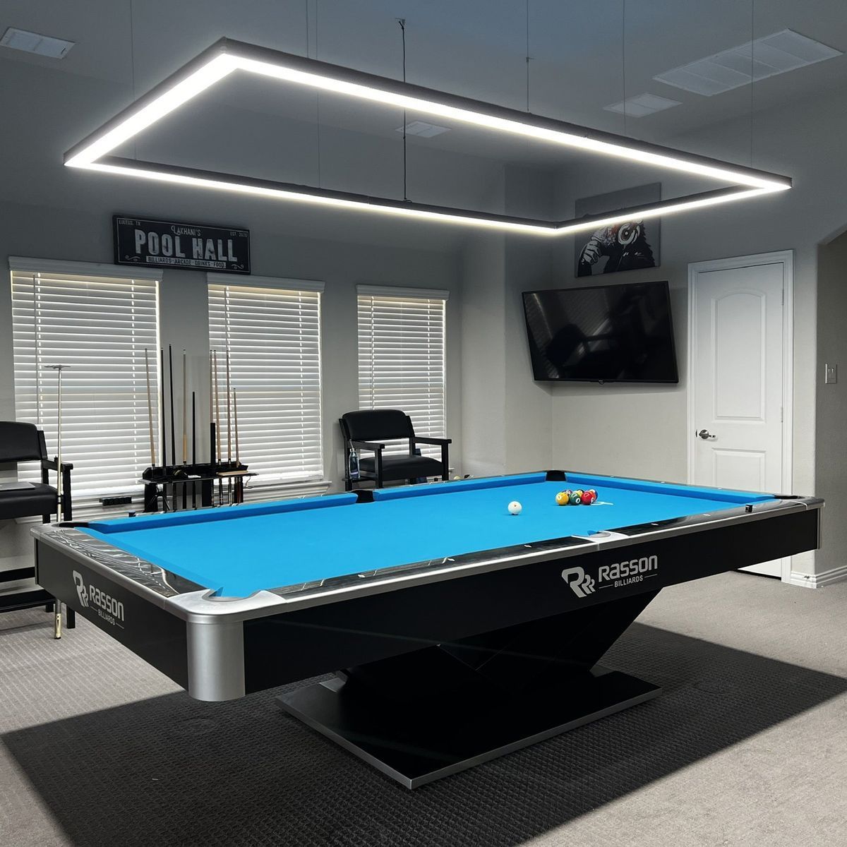 A modern pool table with bright blue felt in a game room, illuminated by a large rectangular overhead light.