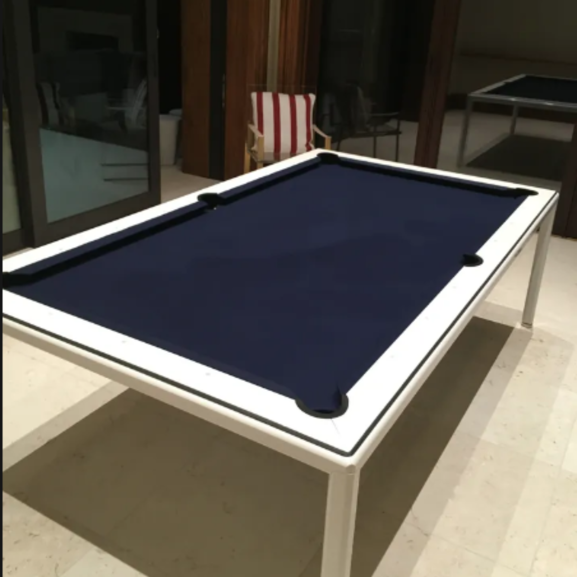 A modern, minimalist pool table with a white frame and deep blue felt, set on a light tiled patio.