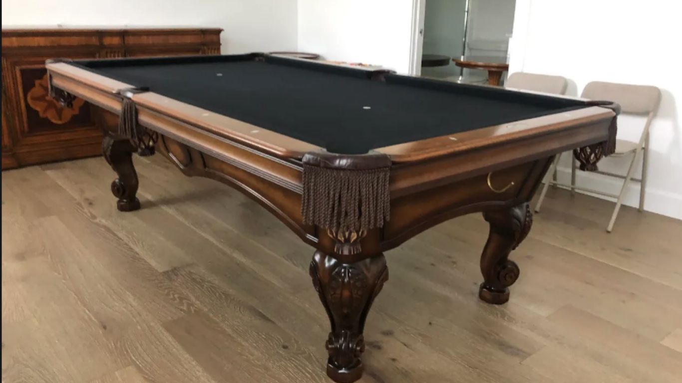 A wooden pool table with a black felt top, intricate carved legs, and fringed pockets, situated in a room with hardwood.