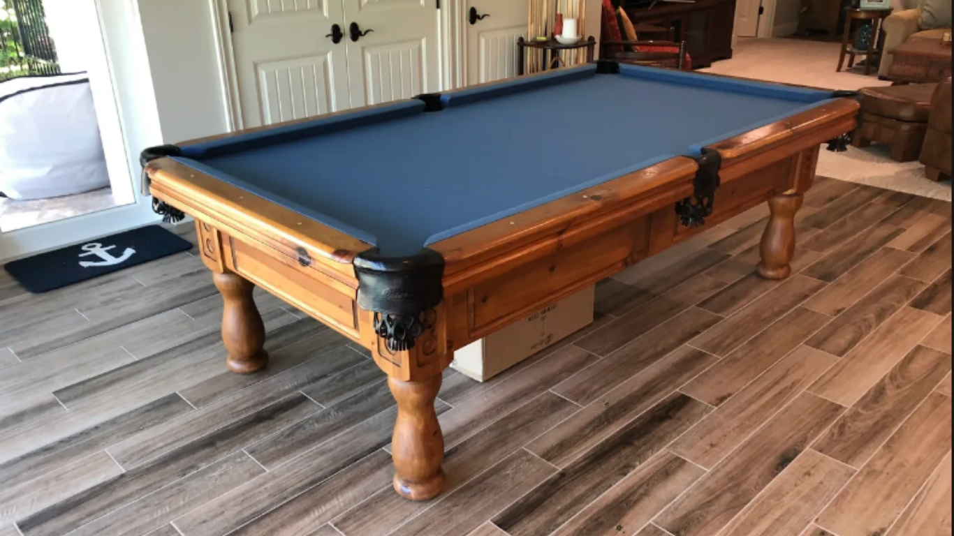 A pool table with a blue felt top and a dark wooden frame sits on a wood-look tile floor in a home interior.