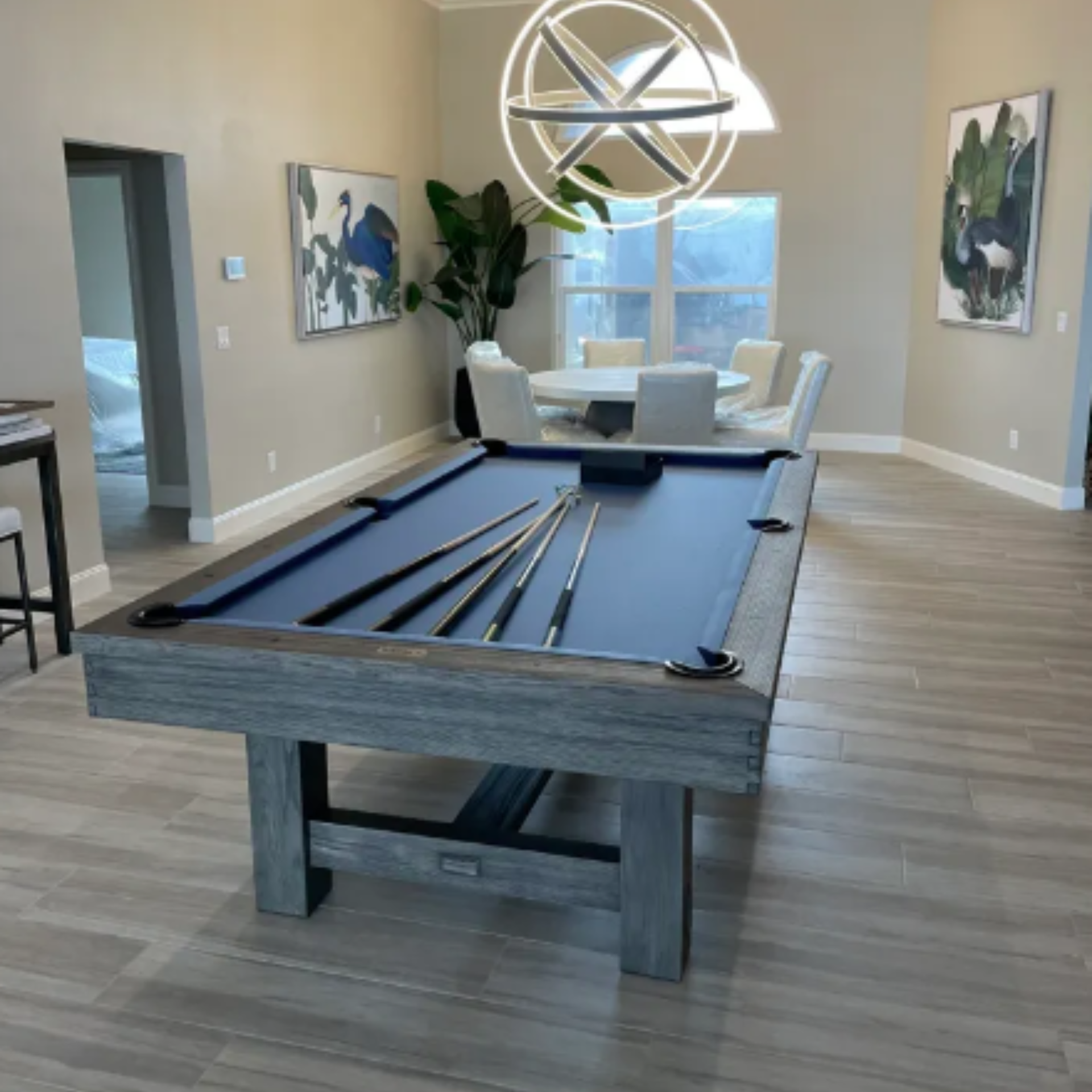 A rustic grey pool table with a blue felt top sits in the foreground of a modern, open-concept room with a dining area.