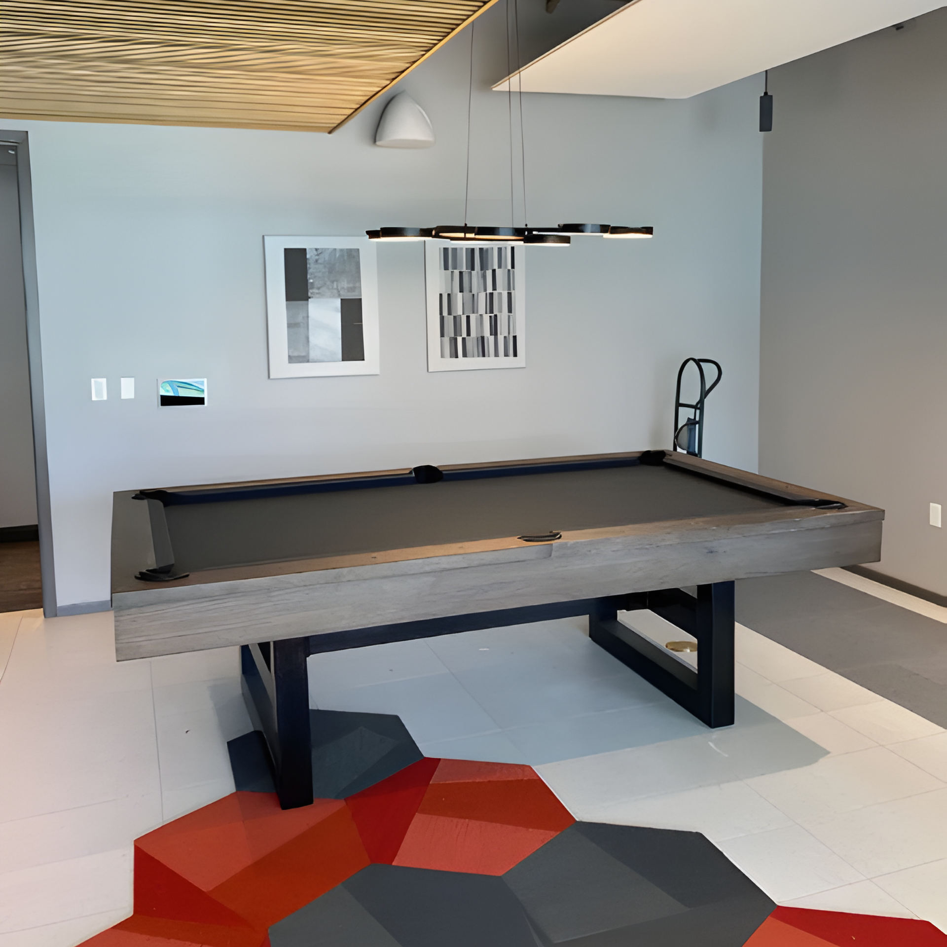 A modern billiard table with a grey felt top and wood-finish rails sits on a geometric red and grey floor in a lounge.