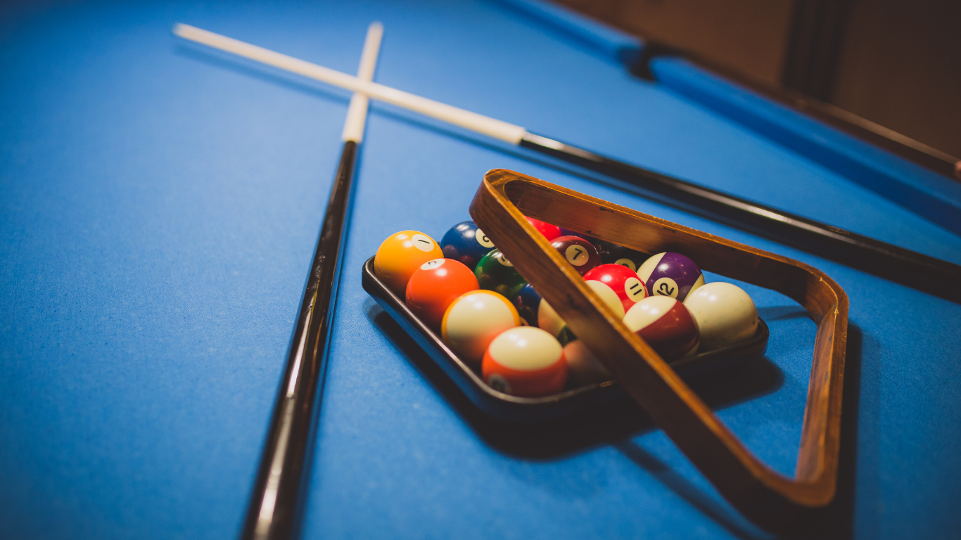 Two pool cues crossed over a blue pool table next to a triangle rack filled with colorful billiard balls.