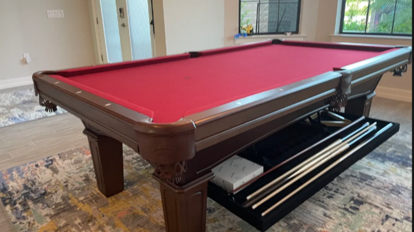 A dark wood pool table with red felt, featuring a built-in storage shelf underneath containing pool cues and accessories.