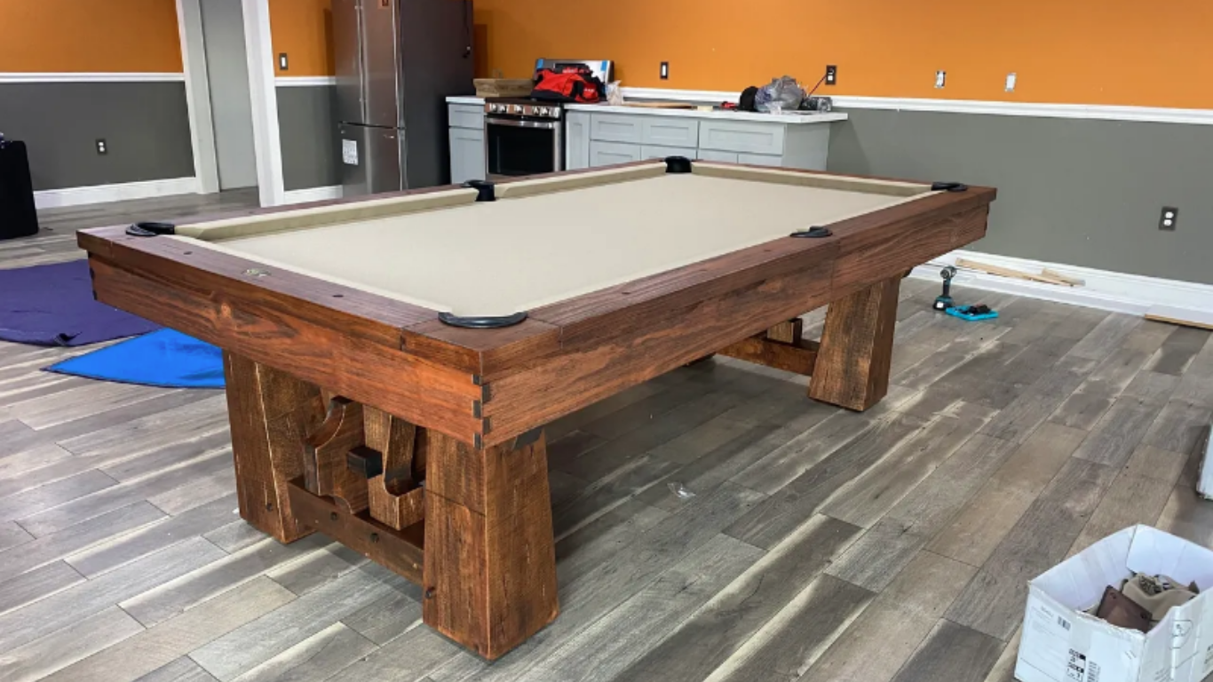 A rustic, dark wood pool table with a tan felt surface sits in a room with orange and gray walls and wood-look flooring.
