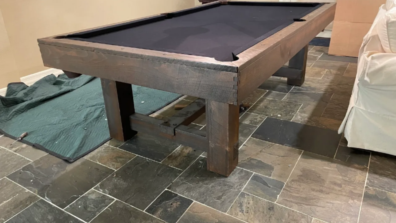 A rustic, dark wood pool table with black felt sits on a dark stone tile floor in an indoor space.