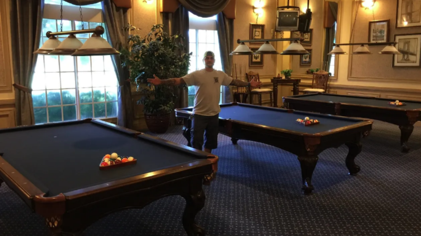 A person with arms outstretched stands between two pool tables in a room with blue carpeting and classic lighting fixtures.