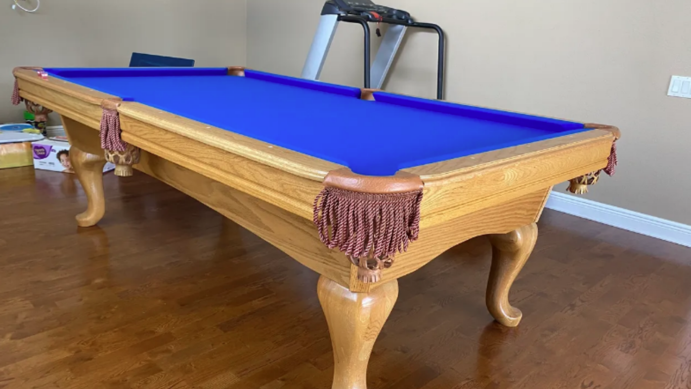 A wooden pool table with a vibrant blue felt surface and decorative fringed pockets, situated in a room with a treadmill.