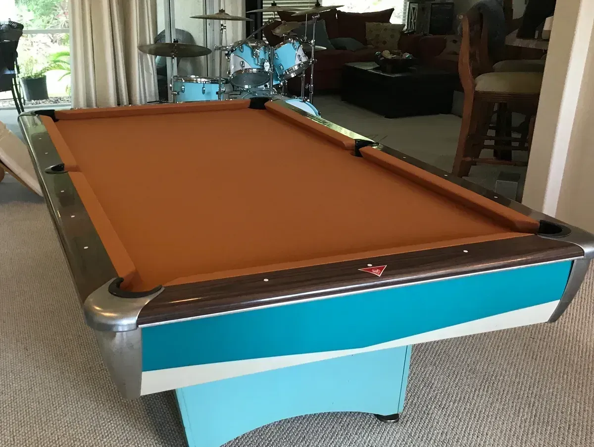 A tan-felted pool table with teal and white side panels, positioned in a room with a drum kit in the background.