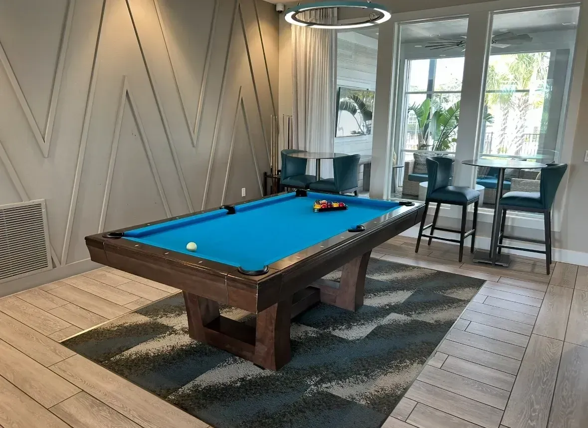 A blue-felt pool table sits on a patterned rug in a room with light wood floors and geometric wall paneling.