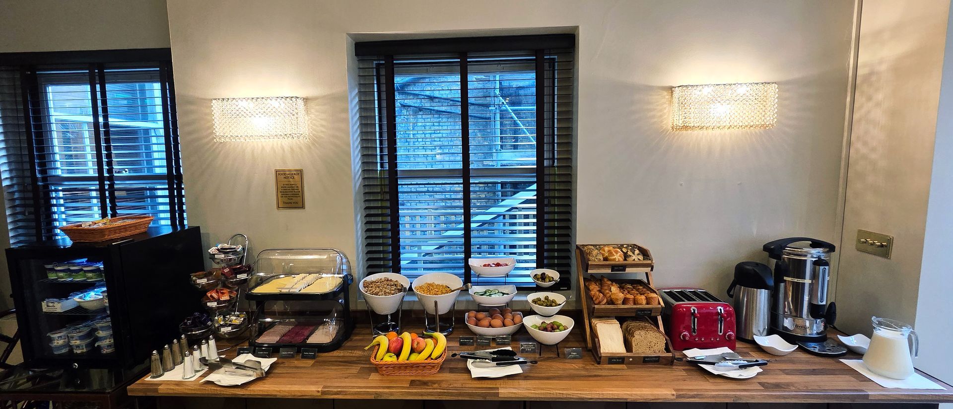 A hotel breakfast buffet spread on a wooden counter with bowls of fruit, pastries, a toaster, and drink dispensers.