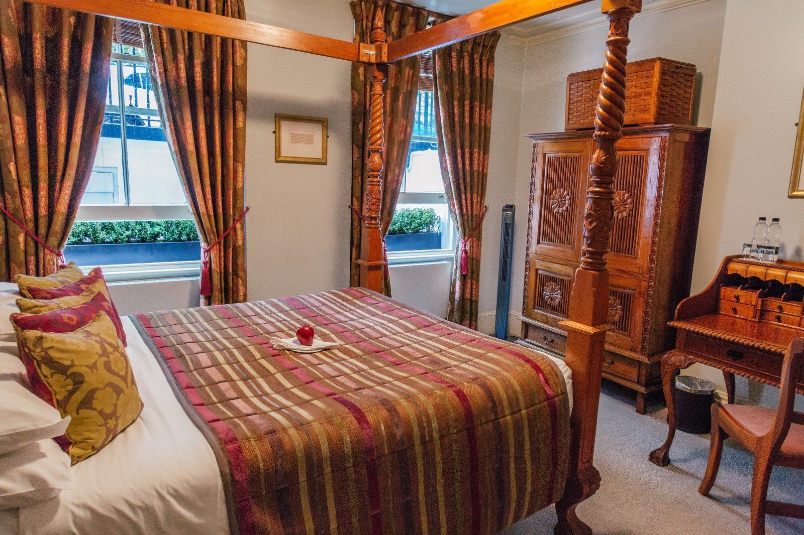 Elegant bedroom with a four-poster bed, wooden wardrobe, and desk; windows with brown curtains.