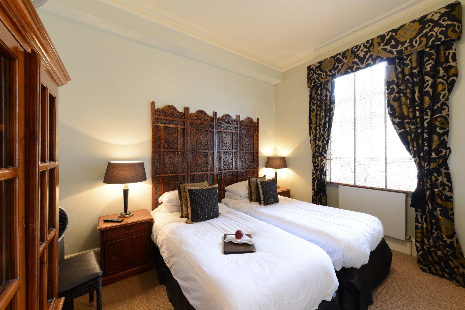 Twin beds in a bedroom with decorative headboard and window with patterned drapes.
