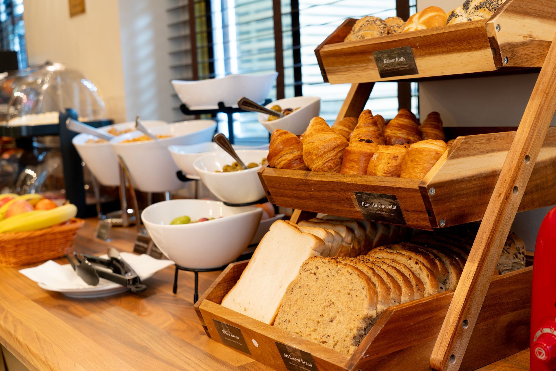 A wooden tiered shelf holding a selection of assorted breads, rolls, and croissants, with bowls of food on the side.