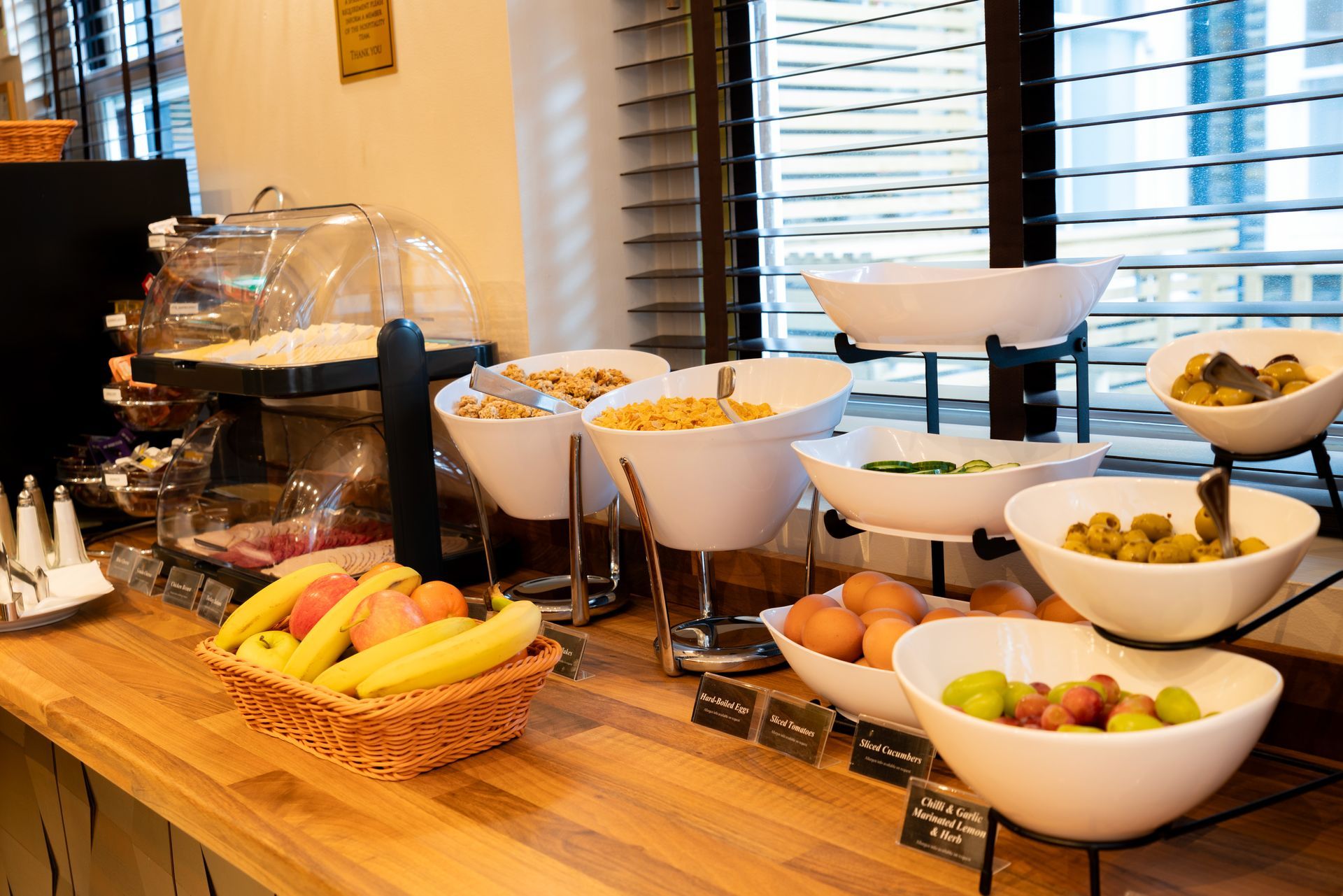 A wooden counter at a breakfast buffet features bowls of fruit, cereal, and hard-boiled eggs with a basket of bananas.