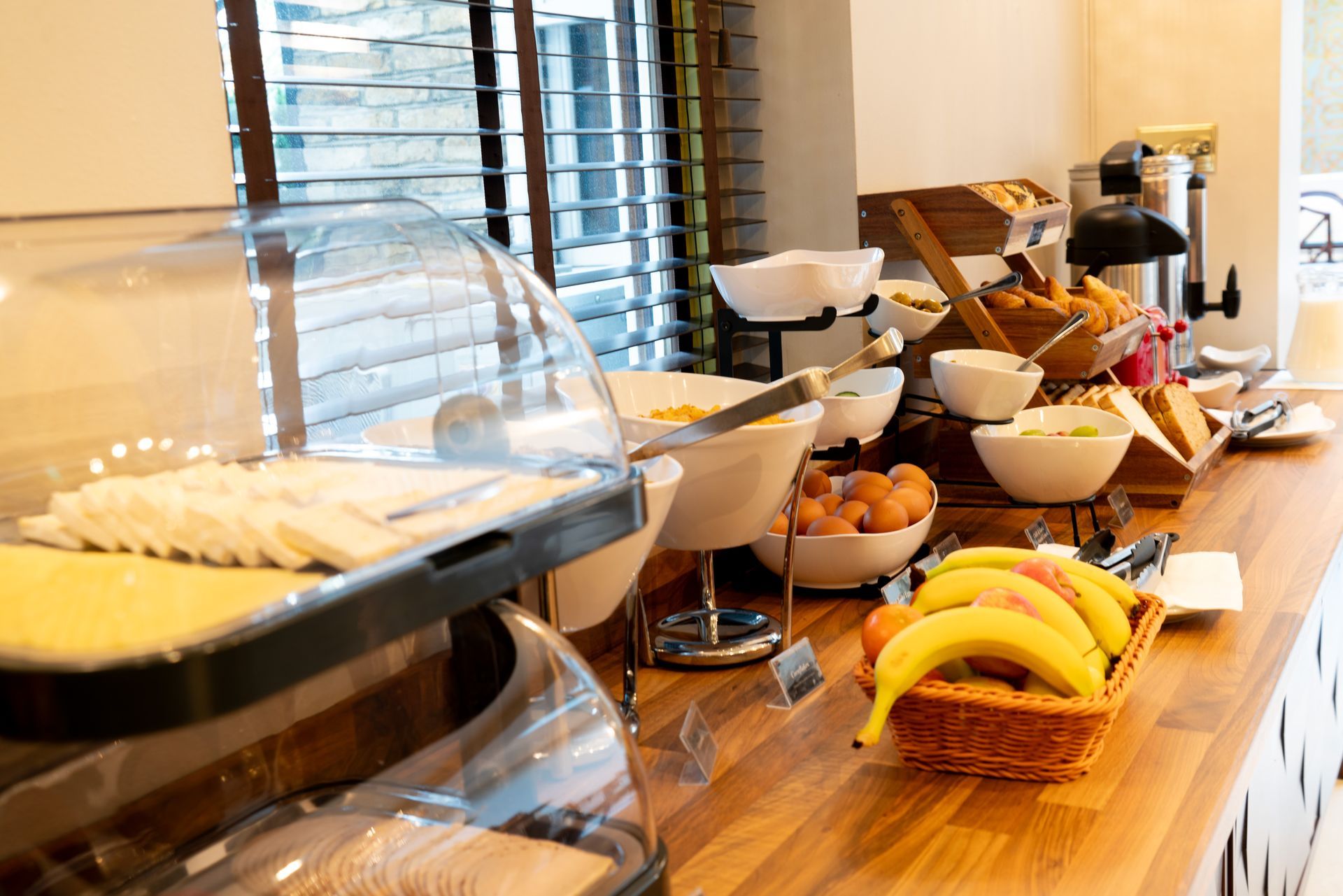 A breakfast buffet display on a wooden counter with bowls of food, fruit, and coffee equipment in a brightly lit room.