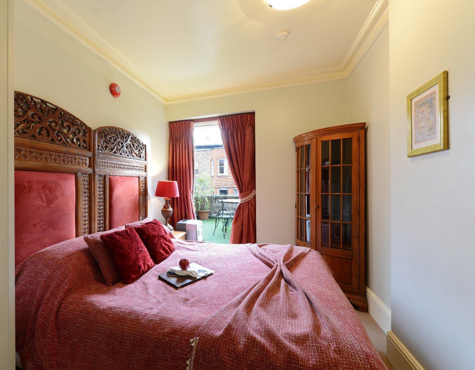 Bedroom with ornate wooden headboard, red bedspread, curtains, and an armoire.