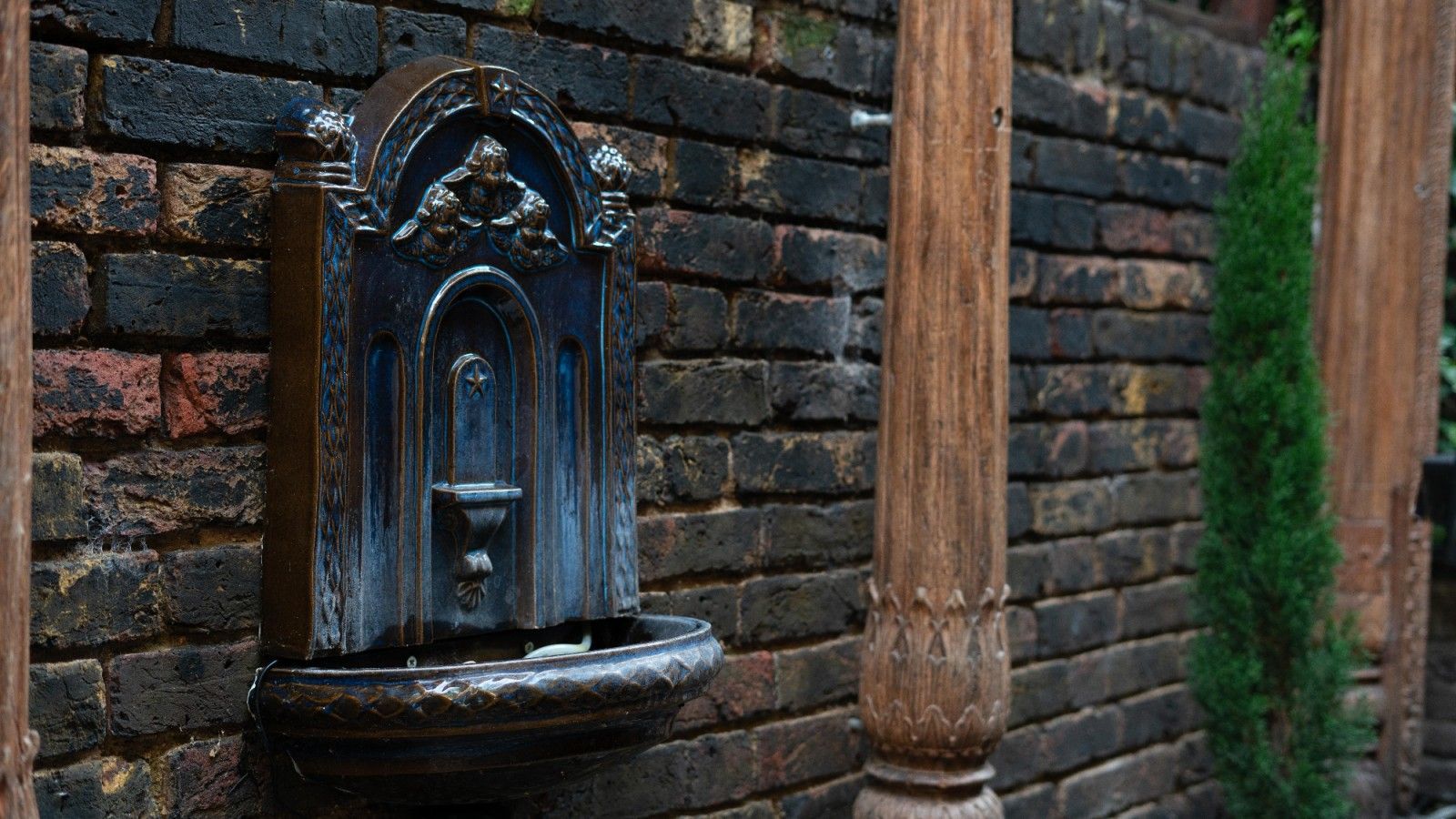 Water fountain on a brick wall, with wooden posts and green bush.