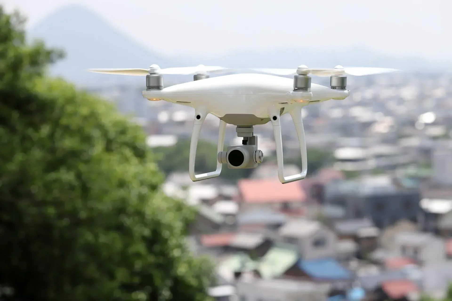 White drone flying over a town with buildings and a mountain in the background.