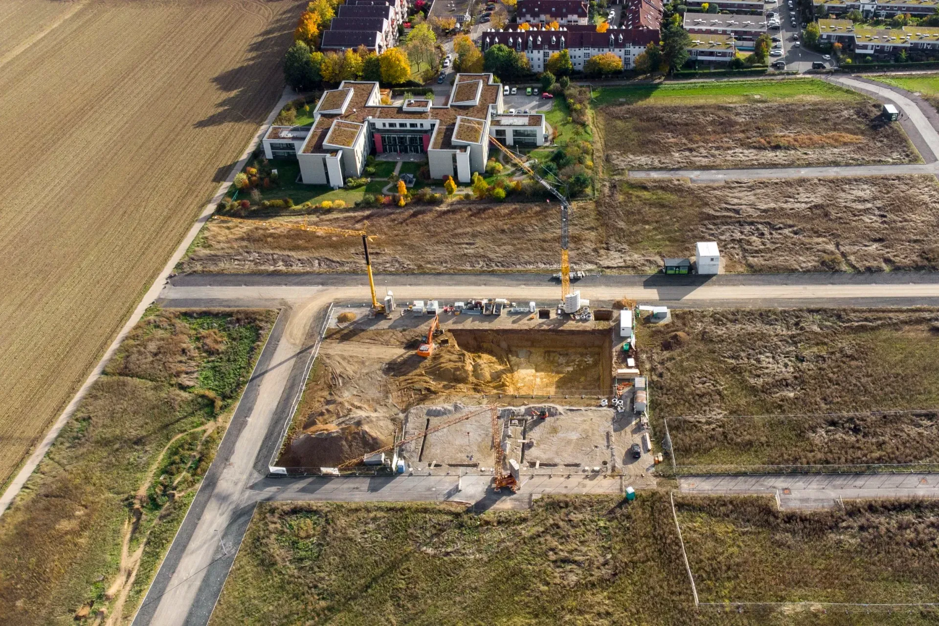 Construction site with earth-moving equipment, a rectangular foundation, and buildings in the background.