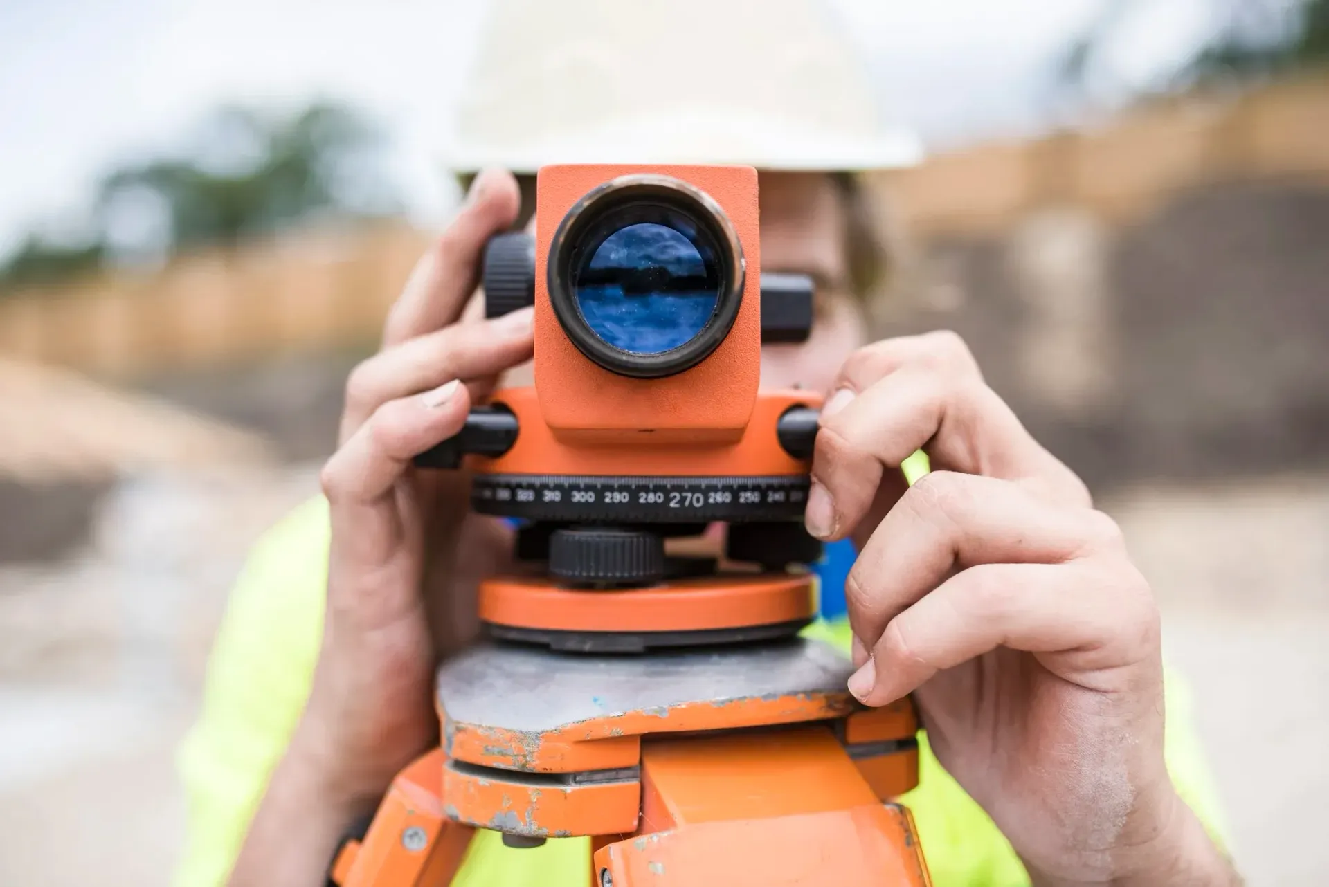 Person in hard hat using orange surveyor's level outdoors.