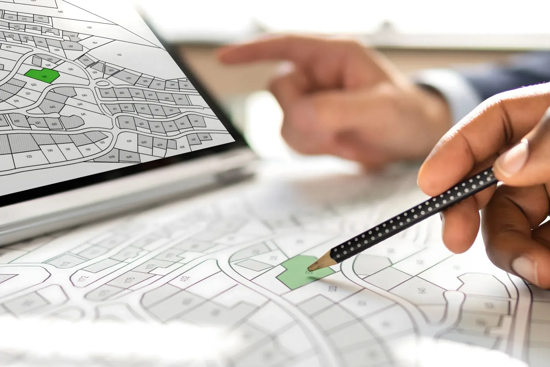 Person pointing a pencil at a highlighted area on a map, with a digital map on a laptop in the background.