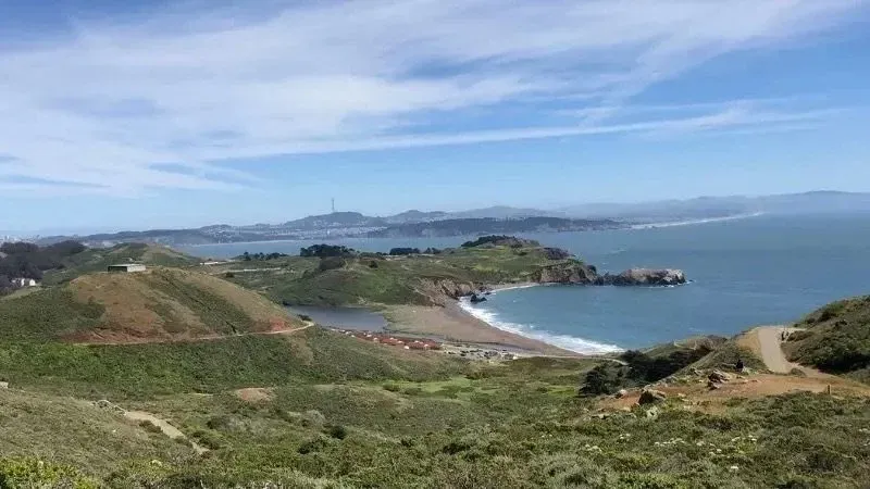 Coastal view: Green hills overlooking a beach, ocean, and distant city with blue sky.