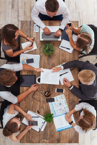 Top-down view of a team meeting around a wooden table with laptops, papers, and coffee cups