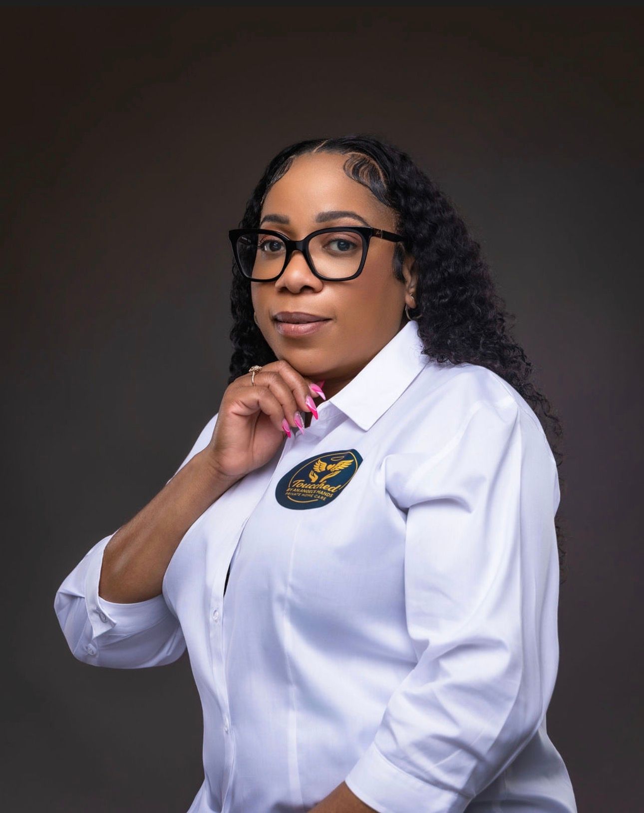 Woman wearing glasses and white shirt with a logo, touching her chin and looking at the camera.