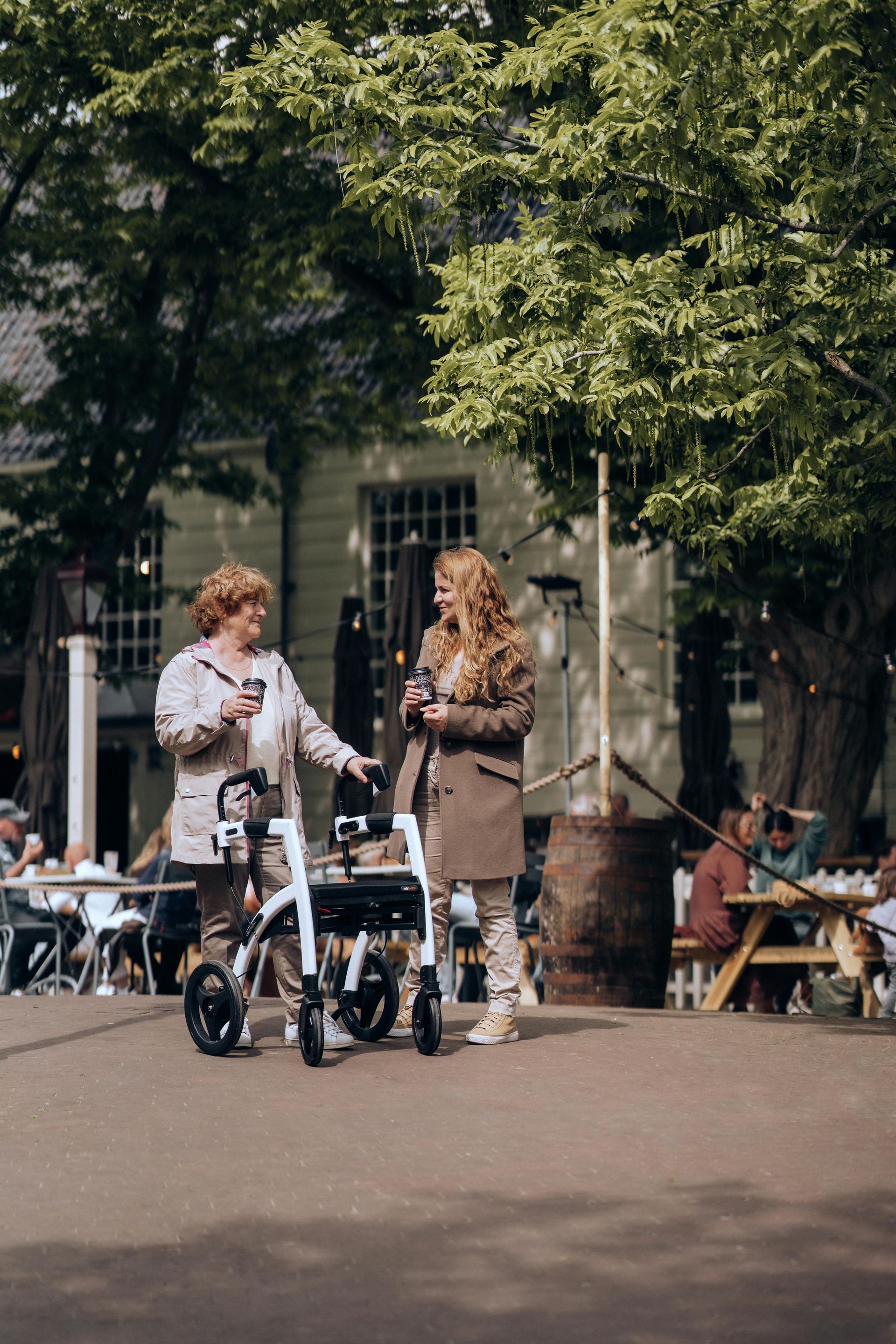 Two women chatting outside, one with a walker. Sunny day, outdoor cafe setting with tables and trees.