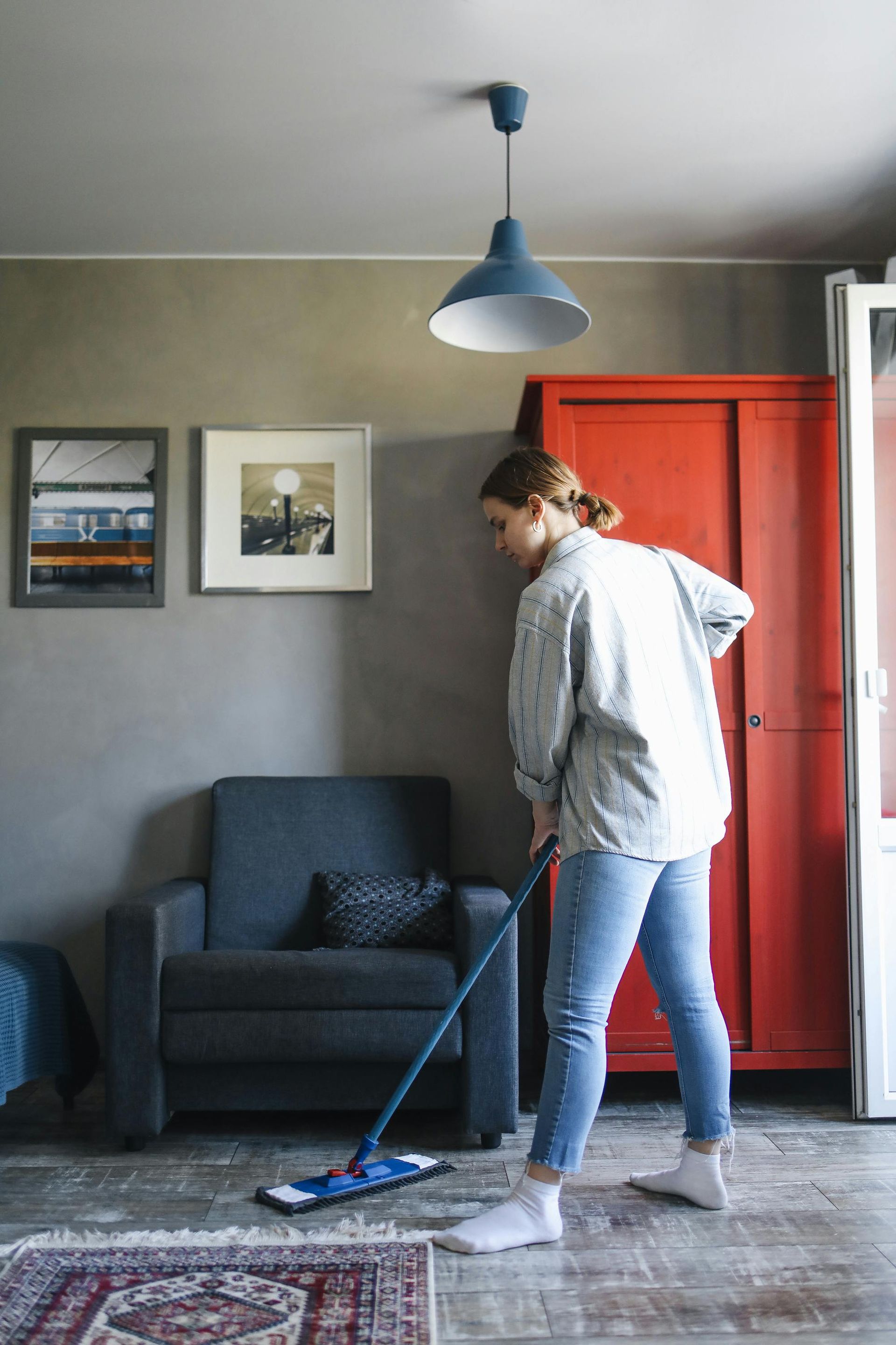 Woman mops a wooden floor in a living room with a red cabinet and a blue armchair.