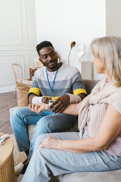 Black man checking the pulse of a seated older woman; both smiling. Indoors, on a sofa.