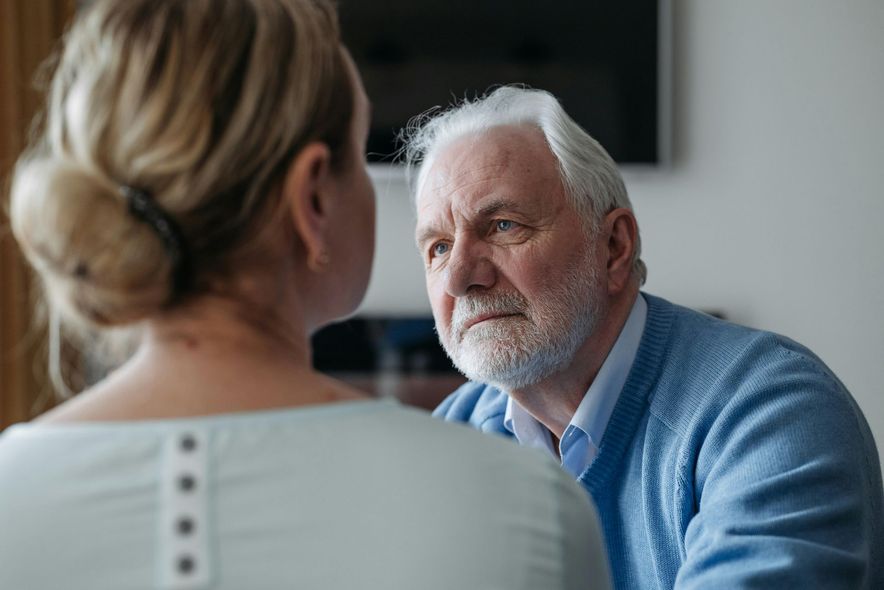 An elderly man gazes intently at a woman; the man is wearing a blue sweater, indoors.