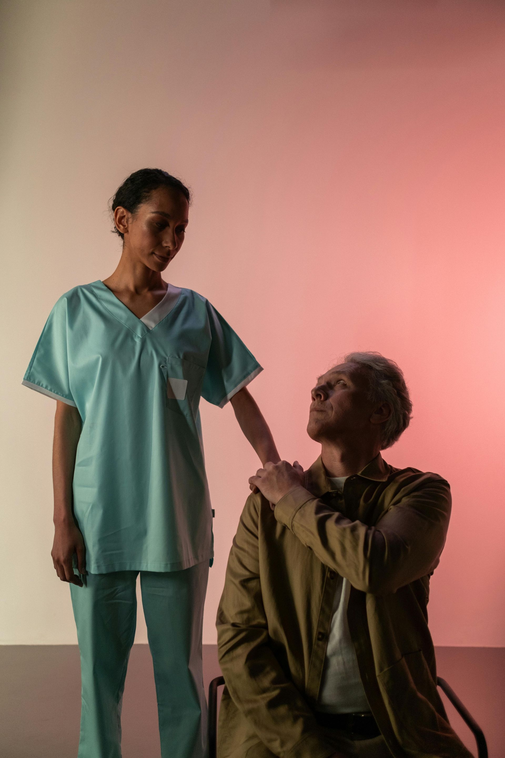 Nurse in scrubs supporting an elderly man in a wheelchair; they are in a brightly lit room.