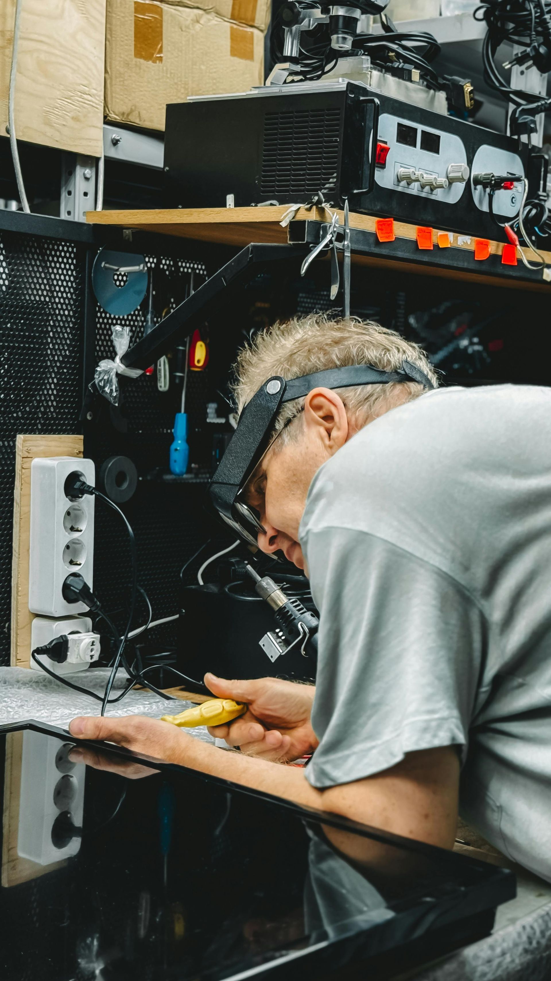 Man working on electronics, wearing a magnifying visor, in a cluttered workshop with equipment.