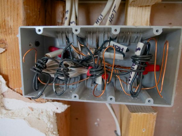 Open gray electrical box with tangled wires, wire nuts, and labels, mounted in a wooden wall.