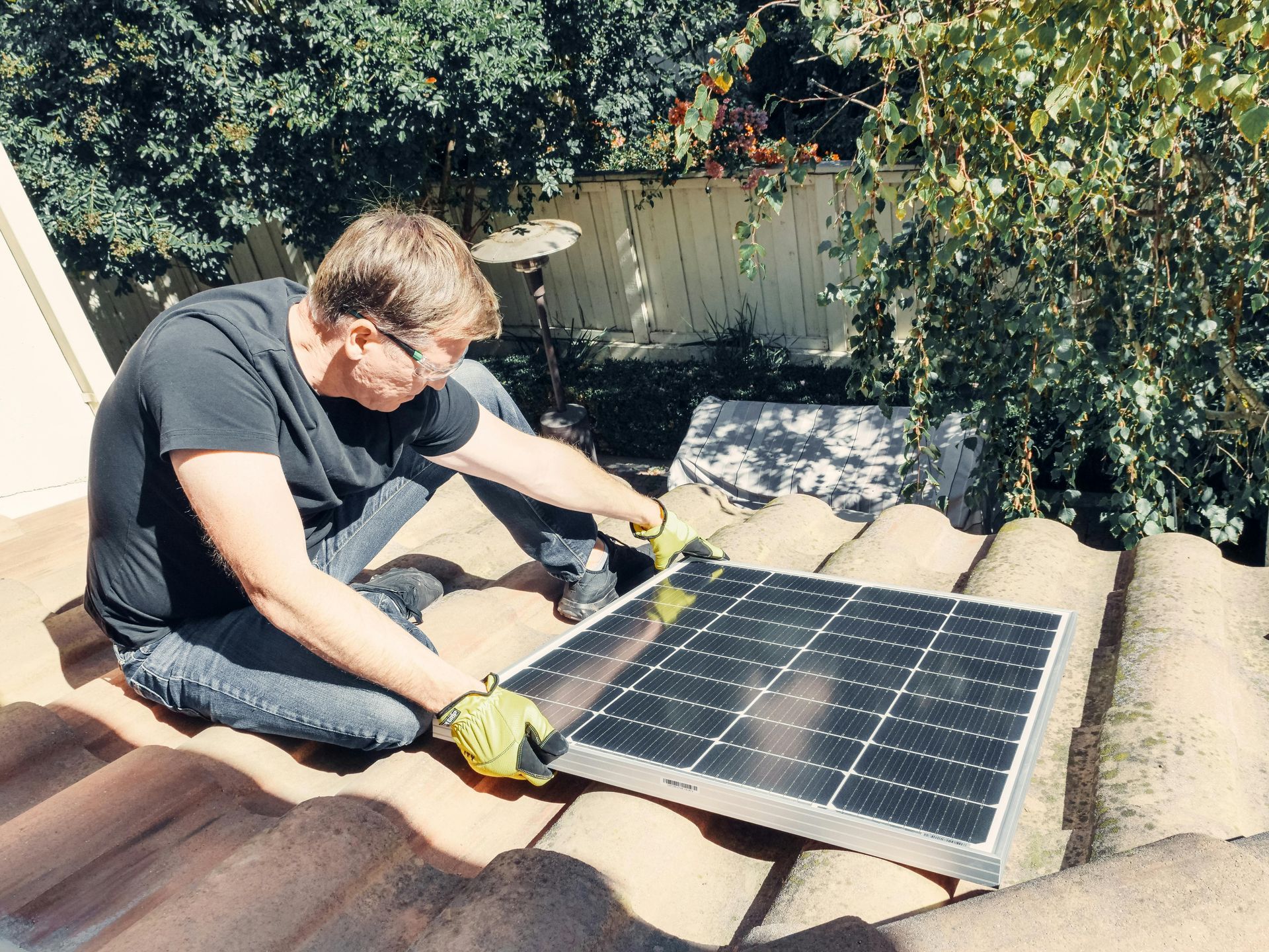 Man installing solar panel on a rooftop. He wears gloves, looking down. Sunny day.